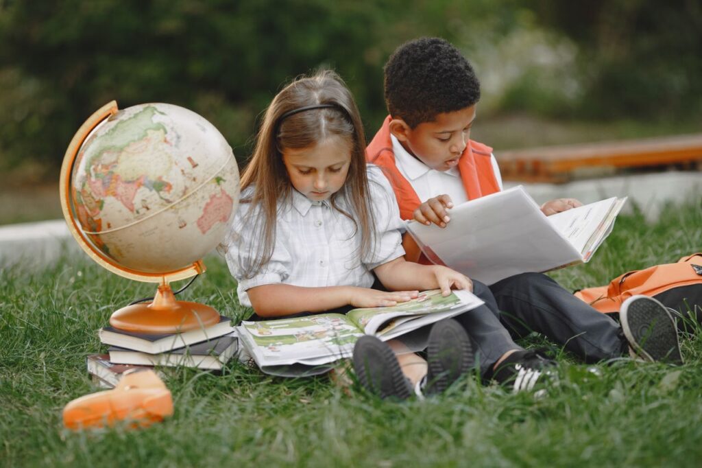 Two children sitting on grass reading books with a globe, promoting education.